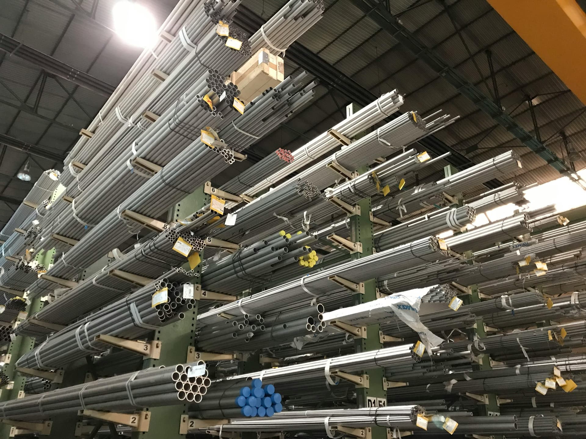 Se observa un estante en un almacén que contiene varios tubos galvanizados apilados horizontalmente. Los tubos están organizados y atados en grupos, mostrando su acabado brillante. El entorno es industrial, con un fondo desenfocado y estanterías visibles.