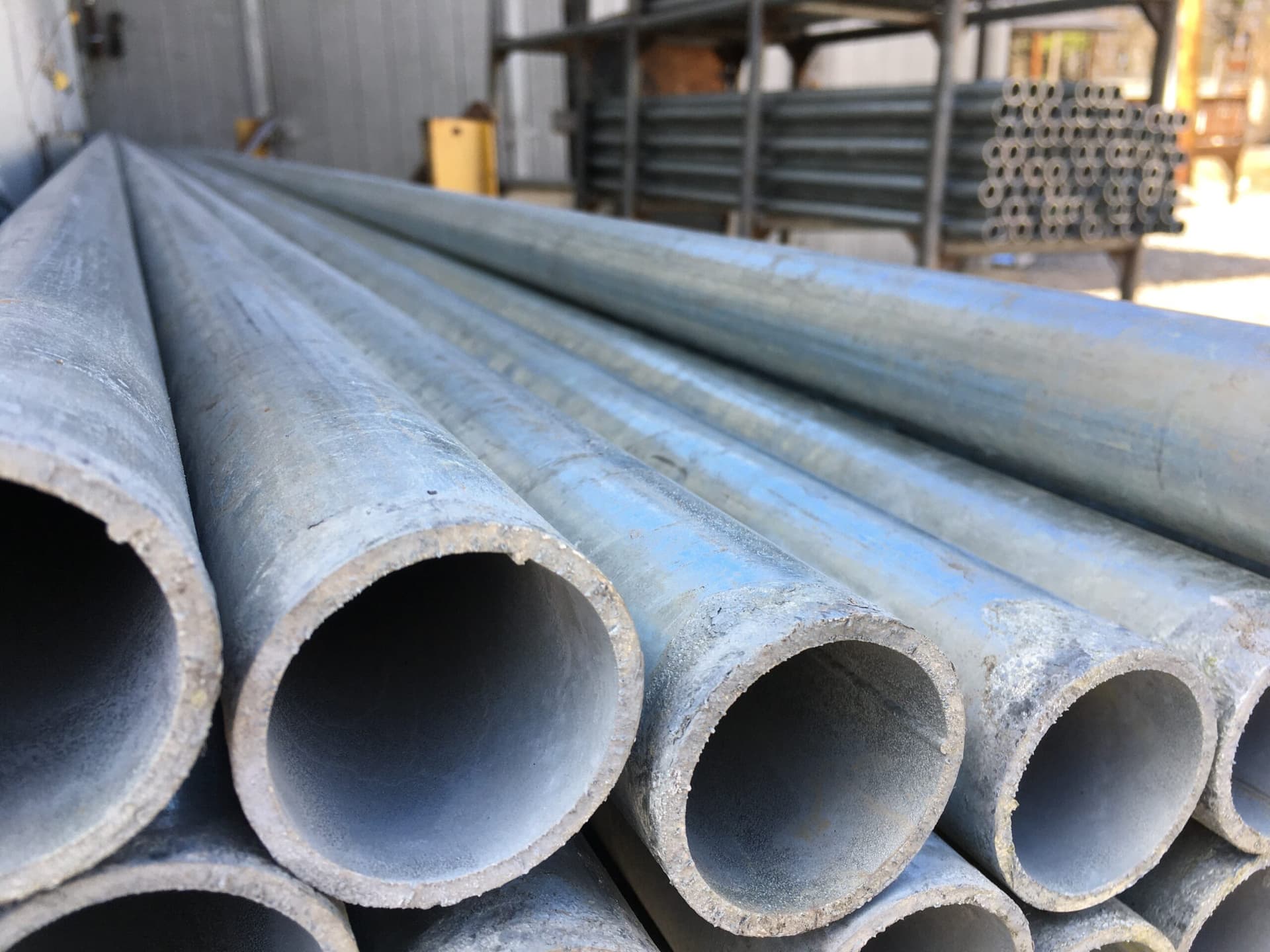 Se observan varios tubos galvanizados apilados, algunos están envueltos con un material transparente y se encuentran sobre una superficie de madera. Los tubos tienen un acabado brillante y se puede ver el interior de uno de ellos. También hay algunos tapones azules visibles en la parte inferior.