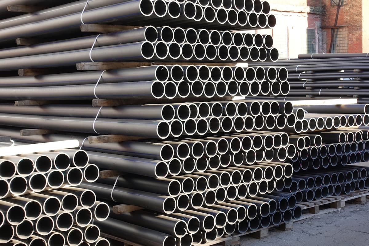 Se observa un gran apilamiento de tubos galvanizados, organizados de manera ordenada. Los tubos son de color negro con extremos visibles y están sujetos con cintas. El fondo muestra un entorno industrial, con estructuras de ladrillo y espacios vacíos.
