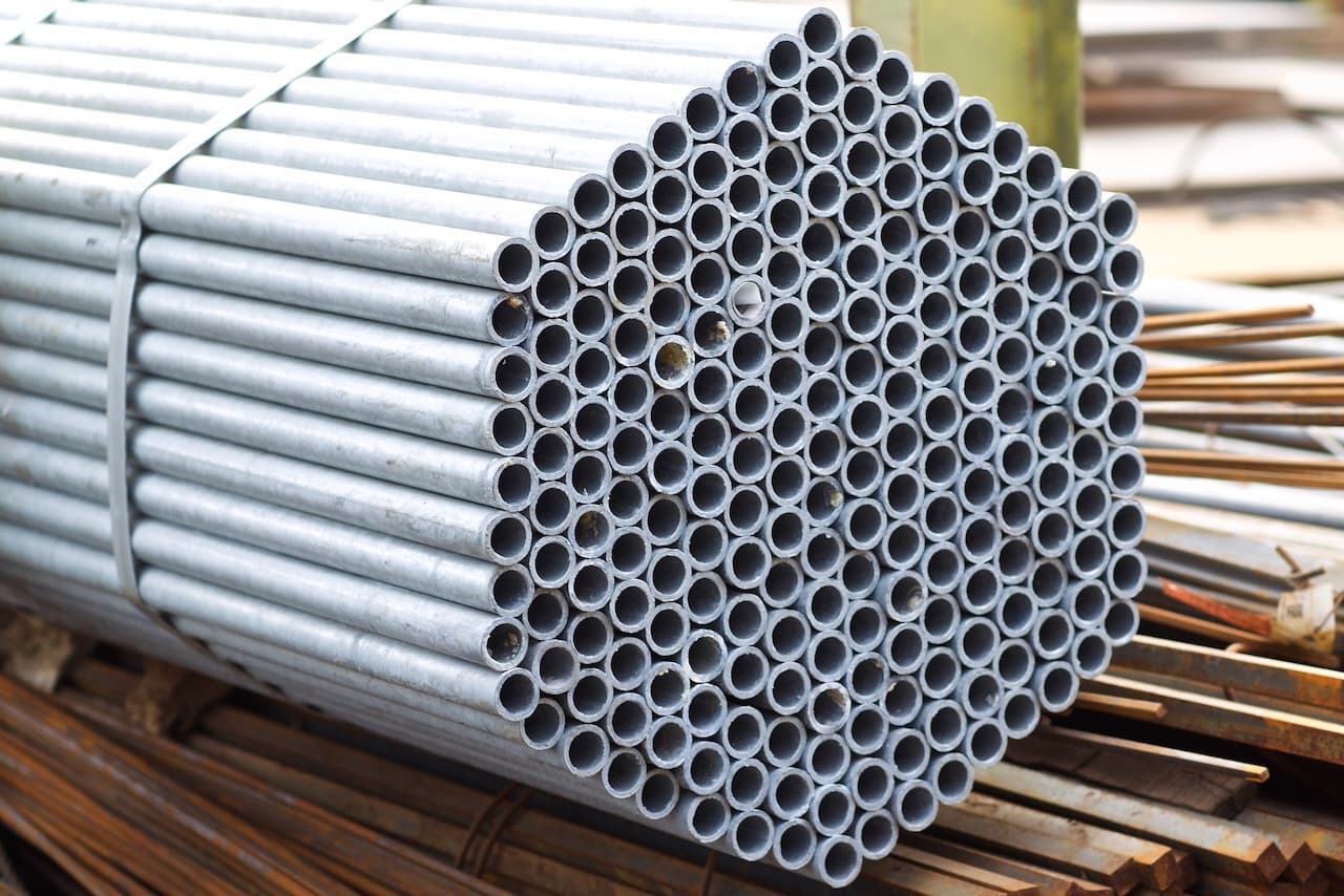 Un grupo de tubos galvanizados apilados de manera ordenada, formando una estructura hexagonal en la parte superior. Los tubos son de color metálico y están colocados sobre una base de madera con otros materiales de construcción visibles en el fondo.