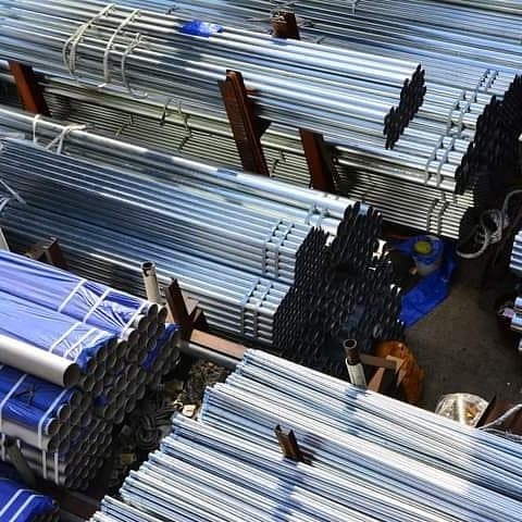 Un conjunto de tubos galvanizados apilados en un área de almacenamiento. Hay tubos de diferentes longitudes, con algunos enrollados en bandas y otros dispuestos de manera ordenada en estanterías. El entorno es industrial con suelo de cemento y algunos objetos adicionales visibles entre los tubos.