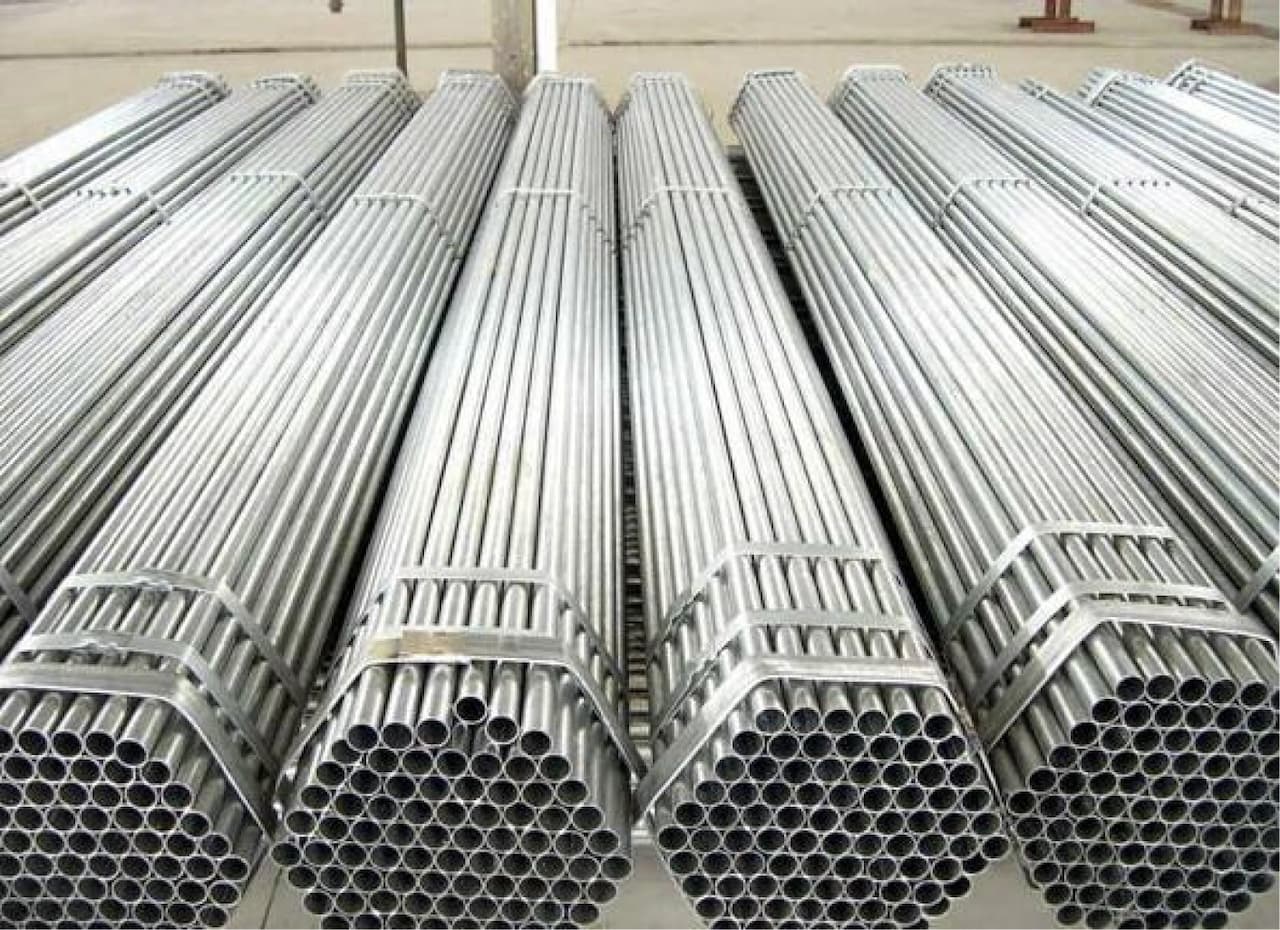Se observan varios tubos galvanizados apilados de manera ordenada, con algunos formando grupos hexagonales en los extremos. La superficie de los tubos tiene un acabado brillante y metálico.
