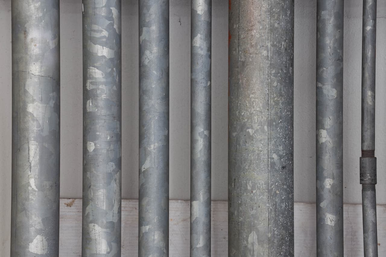 Varios tubos galvanizados dispuestos en una pared, mostrando superficies con un acabado metálico desgastado y marcas visibles. Hay una ligera variación en el grosor y la longitud de los tubos.