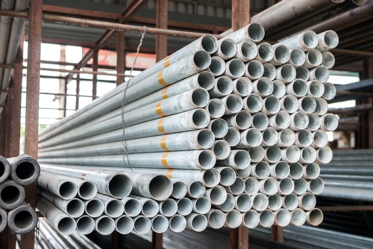 Se observan varios tubos galvanizados apilados en un entorno industrial, organizados en filas y con algunos marcados con bandas de color. El fondo muestra más tubos y estructuras de soporte.