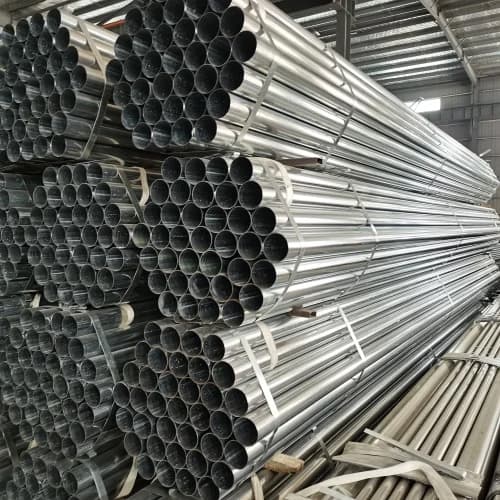 Se observa una pila de tubos galvanizados apilados de manera ordenada en un espacio industrial. Los tubos tienen un acabado brillante y están organizados en filas, algunos son más cortos y otros más largos, todos de diferentes diámetros. El ambiente indica un lugar de almacenamiento o fabricación.