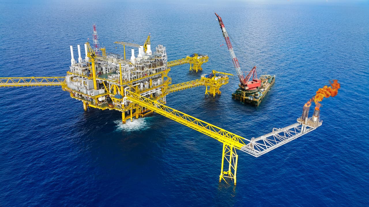 Plataforma marina de extracción de petróleo con estructura amarilla, conectada por pasarelas a otros módulos y un barco. Se observan chimeneas con llamas y el mar azul de fondo.