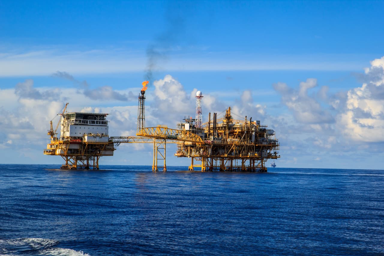 Una plataforma petrolera en el mar, con estructuras metálicas amarillas y varias chimeneas. En una de las chimeneas se observa una llama. El cielo es azul con algunas nubes. El agua está calma.