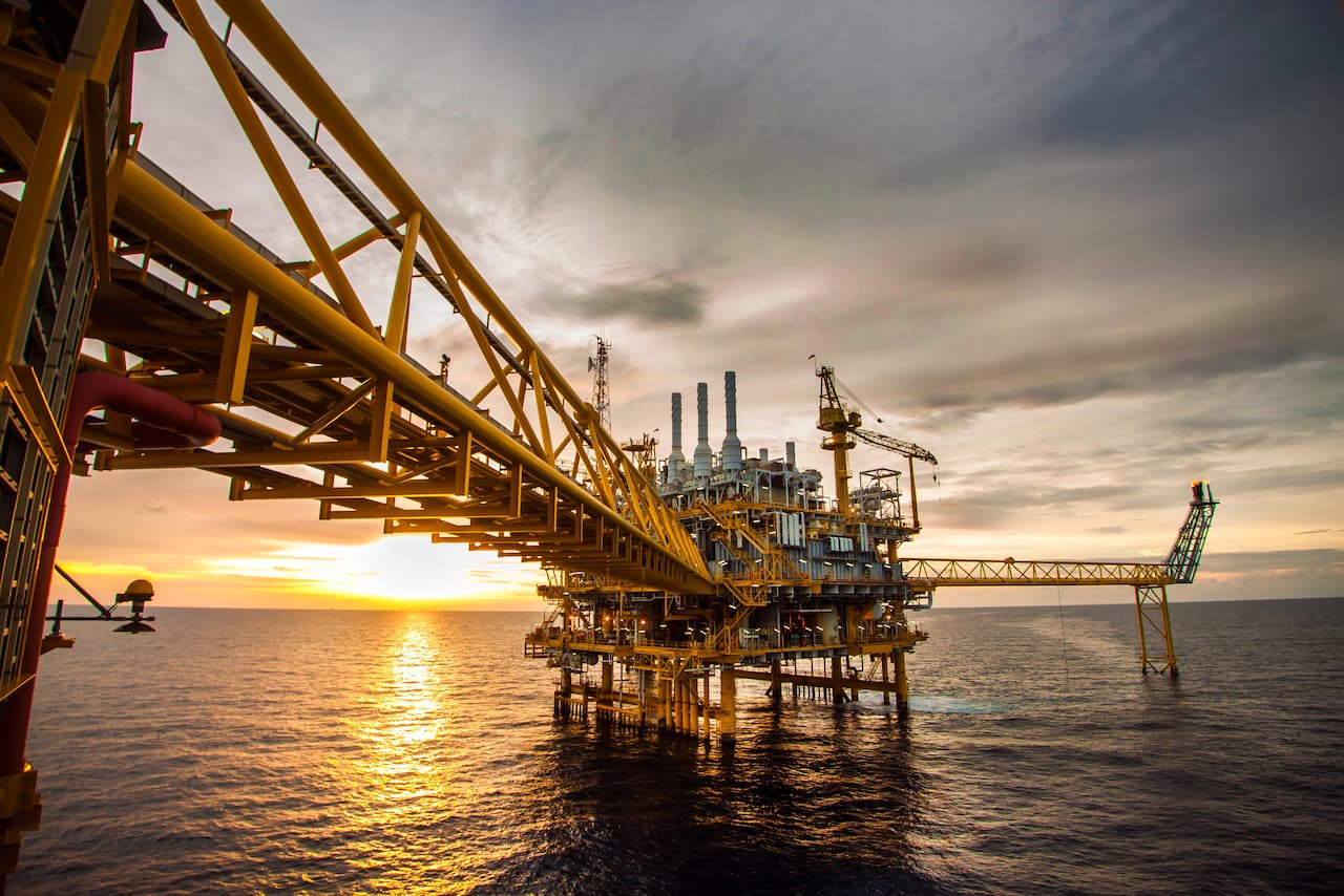 Estructura de una plataforma petrolera en medio del océano, con Tubería Galvanizada conectando diferentes secciones y un atardecer de fondo. Se aprecian chimeneas y maquinaria en la plataforma, con un ambiente de trabajo industrial.